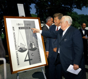 KN-C18708, 31 August 1961 President John F. Kennedy and Senator Alexander Wiley of Wisconsin look at an image of the commemorative stamp created to mark the fiftieth anniversary of the first state Workmen's Compensation Law, enacted in Wisconsin in 1911. The stamp was displayed during a ceremony in honor of the anniversary, held on the South Lawn of the White House, Washington, D.C. Vice President Lyndon B. Johnson stands behind President Kennedy. Please credit “Robert Knudsen. White House Photographs. John F. Kennedy Presidential Library and Museum, Boston”